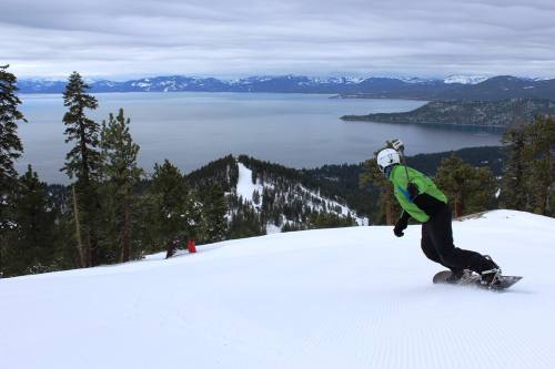 A snowboarder glides down a snowy slope with a scenic view of a lake and mountains in the background.