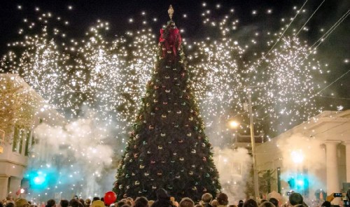 A large Christmas tree adorned with decorations, surrounded by a crowd and illuminated by fireworks at night.