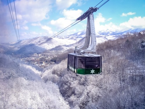 A scenic cable car glides over a snowy landscape with mountains and a small town below, under a blue sky.