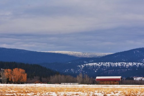 A snowy landscape with a red barn in the foreground and mountains covered in trees in the background under a cloudy sky.