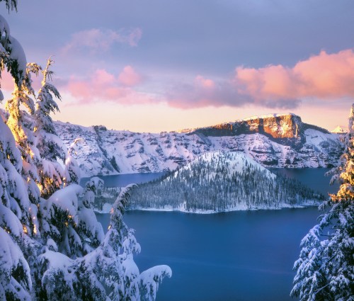 A snowy landscape of Crater Lake, with trees in the foreground and colorful clouds above the serene blue water.
