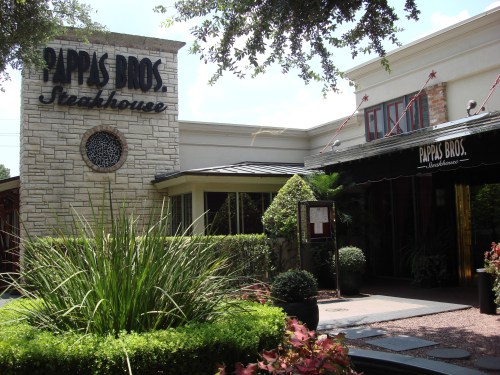 Exterior view of Pappas Bros. Steakhouse, featuring a stone sign, lush landscaping, and a welcoming entrance.