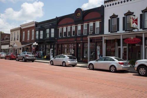 Historic buildings line a brick street, with parked cars and a clear blue sky overhead.