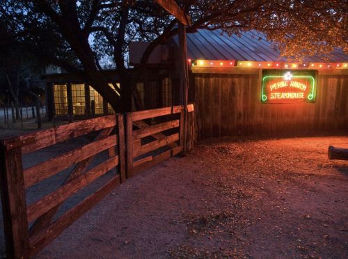A rustic steakhouse sign illuminated at dusk, with a wooden fence and warm lights in the background.