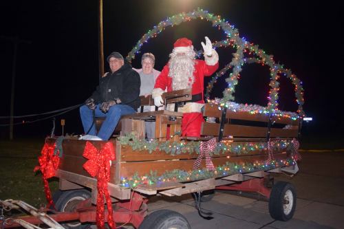 A festive wagon decorated with lights, featuring Santa and two people waving, set against a night backdrop.