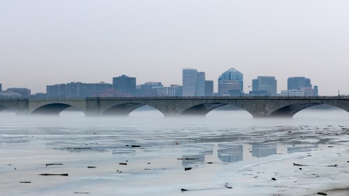 Foggy view of a city skyline behind a stone bridge, with reflections on a calm, icy river surface.