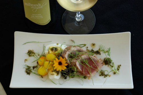 A gourmet dish featuring sliced meat, vibrant garnishes, and a glass of white wine on a black tablecloth.