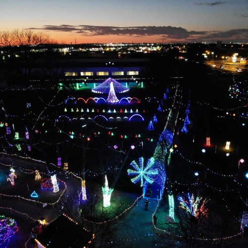 Aerial view of a festive park illuminated with colorful holiday lights against a sunset sky.