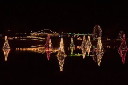 Colorful holiday lights reflect on a calm lake, featuring illuminated trees and festive decorations against a dark sky.