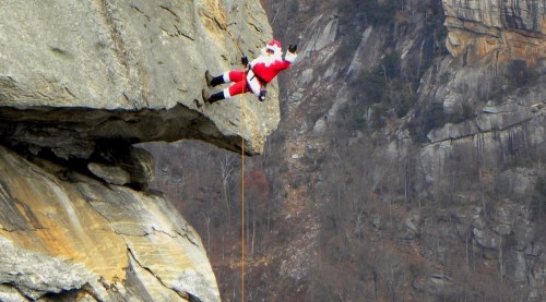 Santa Claus rappelling down a rocky cliff, dressed in his traditional red suit against a mountainous backdrop.