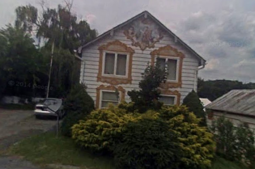 A two-story house with decorative trim, surrounded by greenery and a cloudy sky in the background.