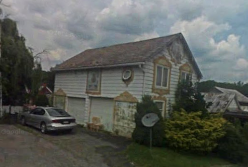 A weathered two-story house with peeling paint, a car parked in the driveway, and a satellite dish in the yard.