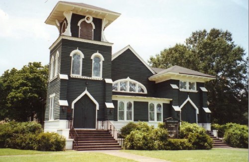A historic building with a tall tower, arched windows, and dark siding, surrounded by greenery and a clear blue sky.
