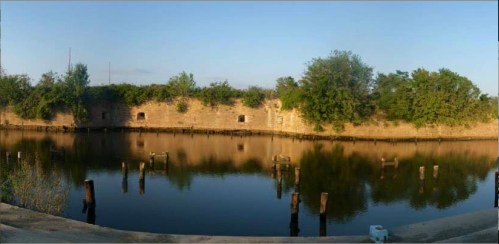 Panoramic view of a calm water body reflecting ruins and greenery along the shore, with wooden posts emerging from the water.
