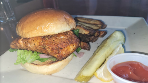 A grilled fish sandwich with lettuce on a bun, served with fries, pickles, and a side of ketchup and lemon.