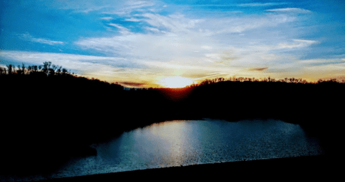 Sunset over a calm lake, with silhouettes of trees and hills against a colorful sky.