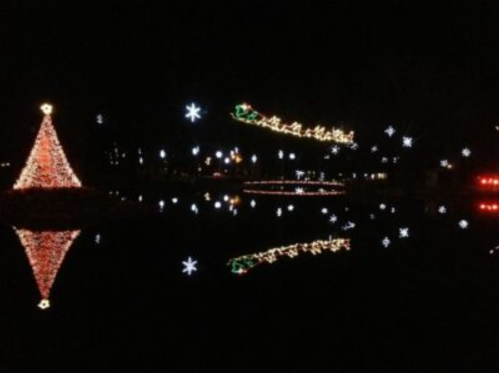 A festive scene at night featuring a lit Christmas tree and a sleigh, reflected in calm water.