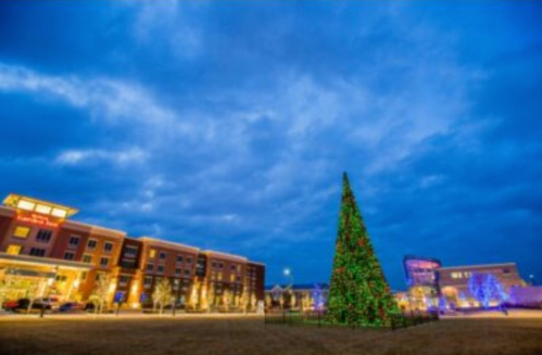 A decorated Christmas tree stands in a park, surrounded by buildings and twinkling lights under a cloudy sky.