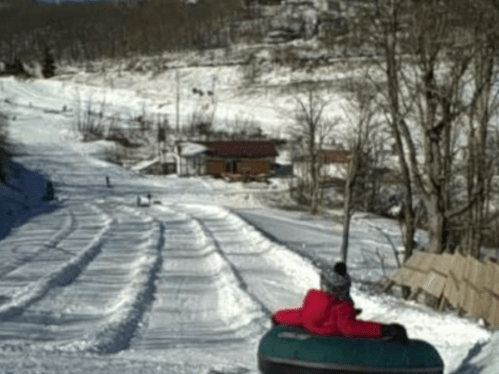 A person sits on a snow tube, ready to slide down a snowy hill with trees and a cabin in the background.