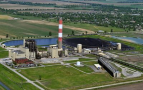Aerial view of a power plant with a tall smokestack, coal storage, and surrounding fields and water bodies.