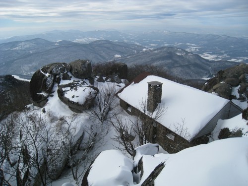 A snow-covered mountain landscape with a small stone cabin surrounded by large boulders and distant hills.