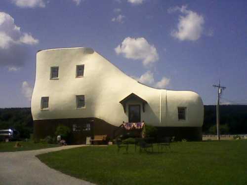 A whimsical shoe-shaped house with a white exterior and brown base, set against a blue sky and green lawn.