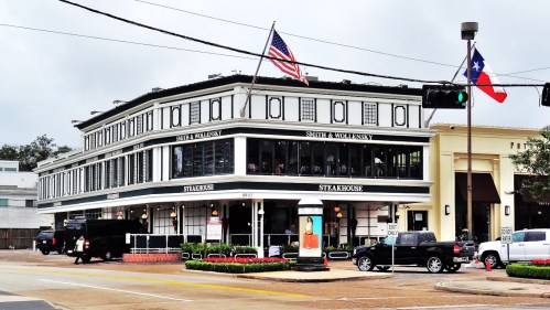 A two-story steakhouse building with an American flag and Texas flag, surrounded by greenery and street signs.