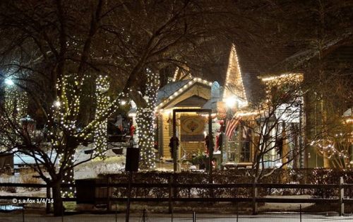 A cozy winter scene featuring a warmly lit house adorned with holiday lights and surrounded by snow-covered trees.
