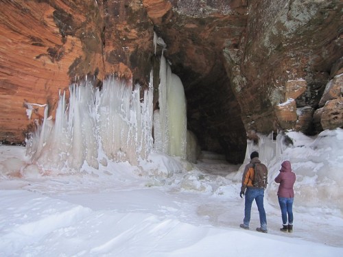 Two people stand in a snowy cave, admiring large ice formations and frozen waterfalls on the rocky walls.