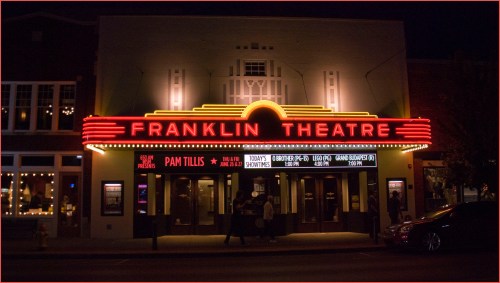The illuminated facade of Franklin Theatre at night, featuring a marquee with event listings and a welcoming entrance.