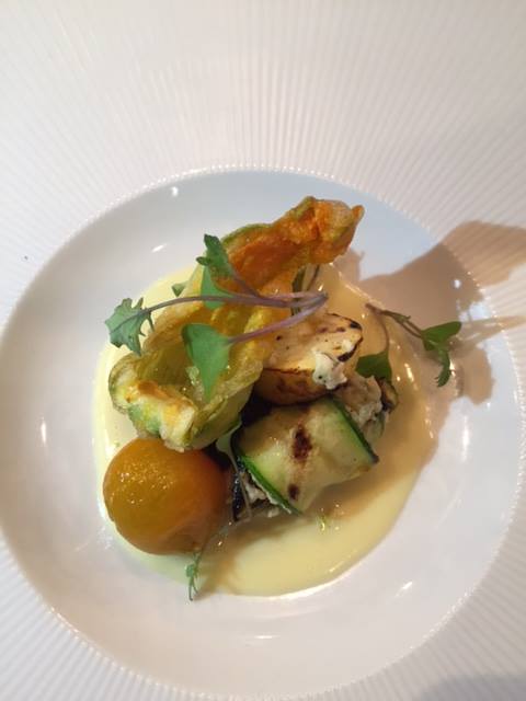 A gourmet dish featuring grilled vegetables, a stuffed flower, and a creamy sauce, elegantly presented on a white plate.
