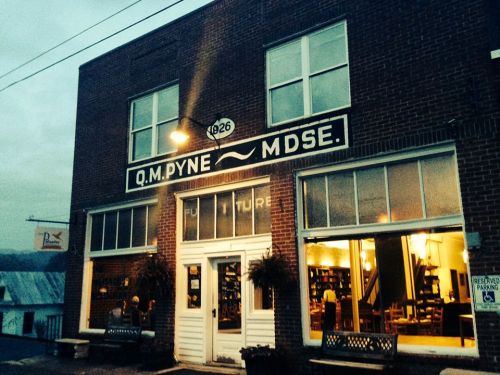 A brick storefront with "Q.M. Pyne MDSE" sign, featuring large windows and warm lighting inside.