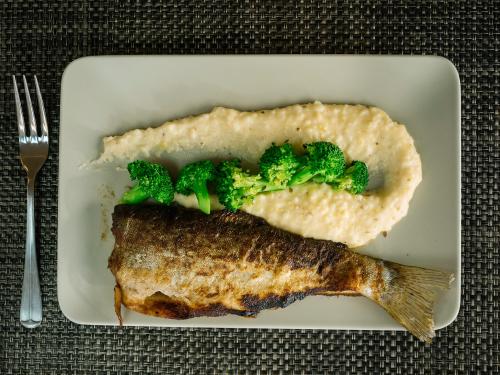 A plate with a whole fish, broccoli, and creamy grits, served on a textured tablecloth.