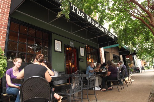Outdoor dining scene at The Wooden Nickel, with patrons enjoying meals at tables under a canopy of trees.