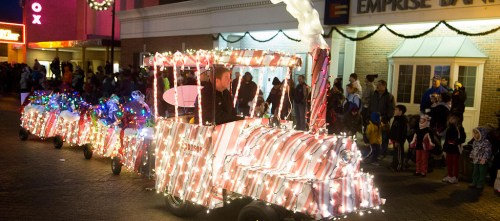 A festive train decorated with lights and candy cane stripes, surrounded by a crowd enjoying a holiday parade.