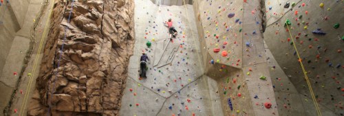 Two climbers ascend a colorful indoor rock climbing wall with various holds and a textured rock surface.