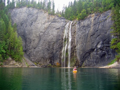 A person in a kayak paddles near a waterfall surrounded by tall trees and rocky cliffs.