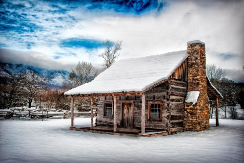 A rustic log cabin covered in snow, surrounded by a winter landscape and mountains under a cloudy blue sky.
