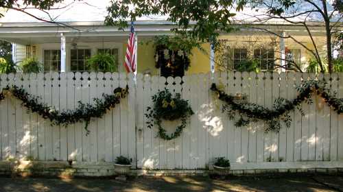 A white picket fence adorned with greenery and wreaths, leading to a house decorated for the holidays. An American flag is visible.