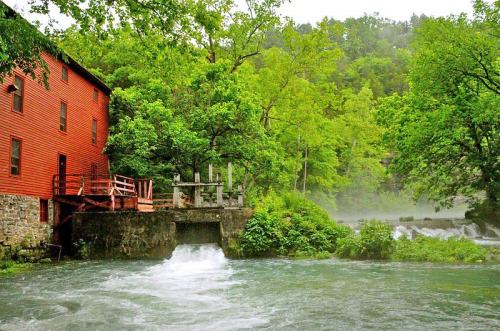 A red mill building beside a flowing river, surrounded by lush green trees and misty hills in the background.