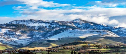 Snow-capped mountains under a blue sky with scattered clouds, overlooking green hills and a small village.
