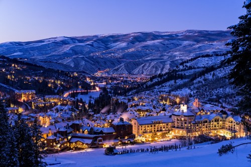A snowy mountain village at dusk, with twinkling lights and a backdrop of snow-covered hills.