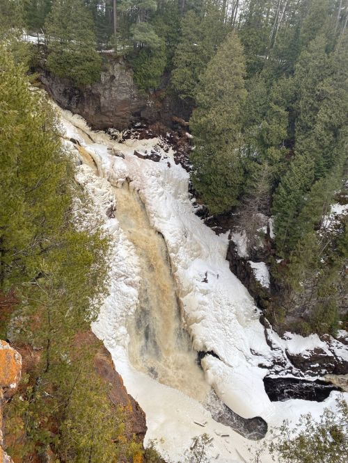 A waterfall cascading over rocky terrain, surrounded by evergreen trees and patches of snow.