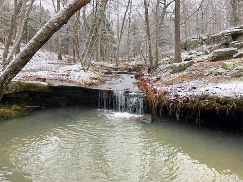 A serene winter scene featuring a small waterfall flowing into a calm pool, surrounded by snow-covered trees and rocks.