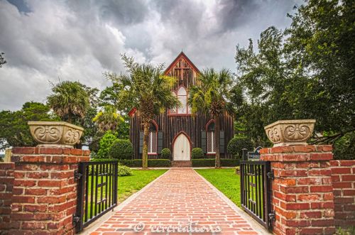 A charming wooden church with a steep roof, surrounded by greenery and palm trees, under a cloudy sky.