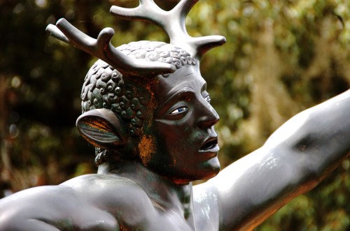 A close-up of a bronze statue depicting a figure with antlers and a muscular build, set against a blurred natural background.