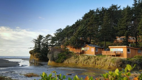 Cozy wooden cabins nestled among trees on a coastal cliff, overlooking a sandy beach and calm waters.