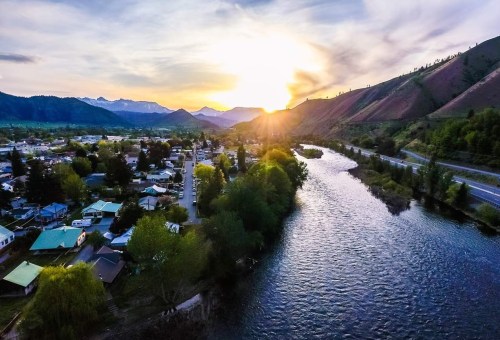 Aerial view of a river winding through a small town, with mountains and a sunset in the background.