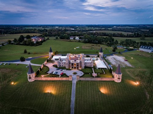 Aerial view of a grand castle with turrets, surrounded by green fields and a winding driveway.