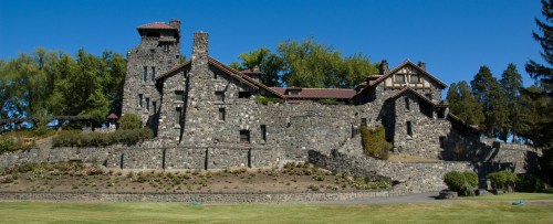 A large stone castle with a tower, surrounded by trees and a grassy area under a clear blue sky.